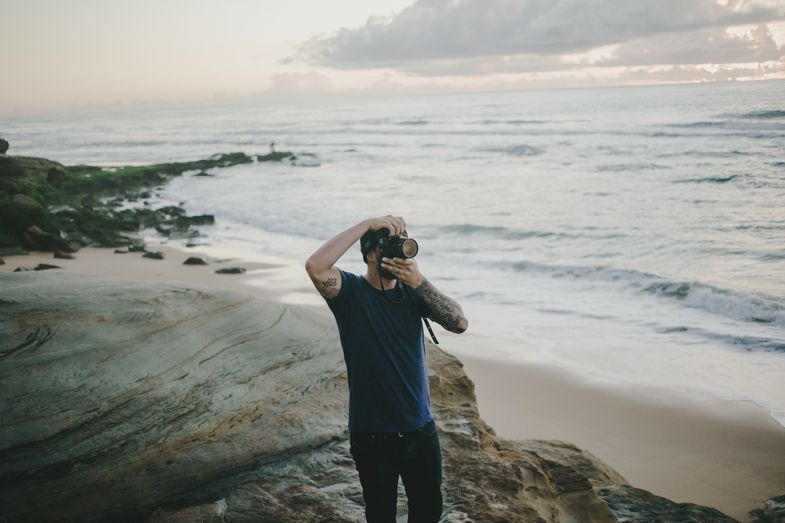 A photographer takes pictures at sunset on a rocky beach overlooking the ocean.