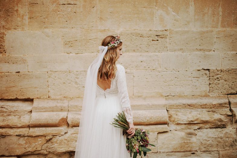 Back view of bride in flowing dress with bouquet against textured stone wall.