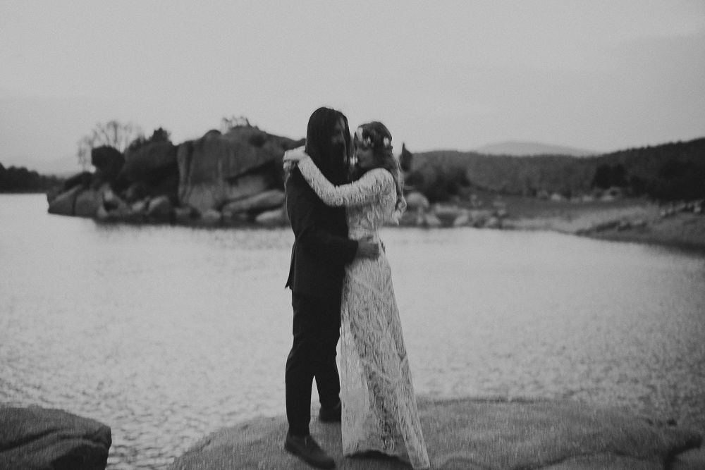 Black and white wedding photograph on lakeside rocks with mountain scenery.