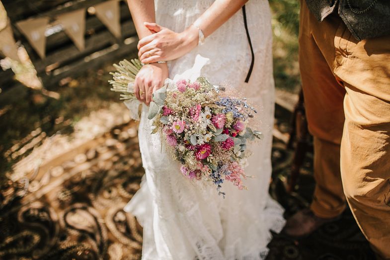 A delicate bouquet of pink and purple wildflowers held against a white lace wedding dress.