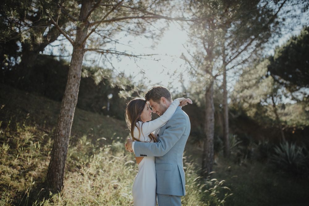 Intimate embrace between couple in formal attire against sunlit forest backdrop.