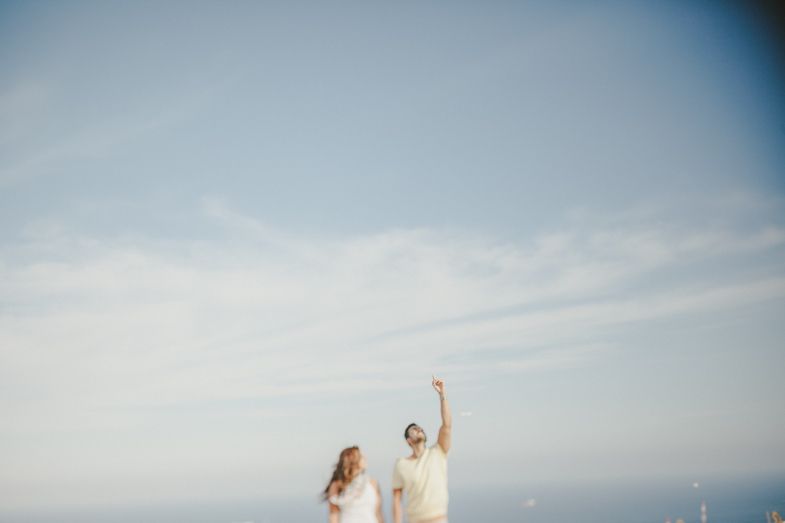 A couple in white clothing stands against a bright sky with their arms raised triumphantly.