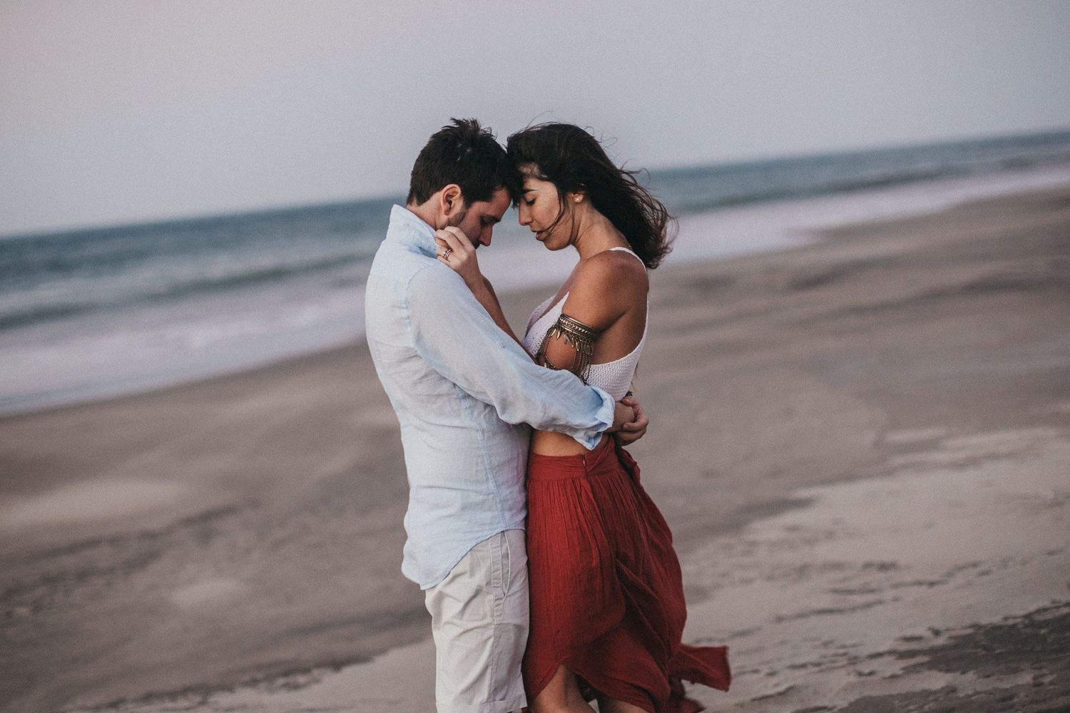 Romantic beach engagement photo with couple embracing in casual attire.
