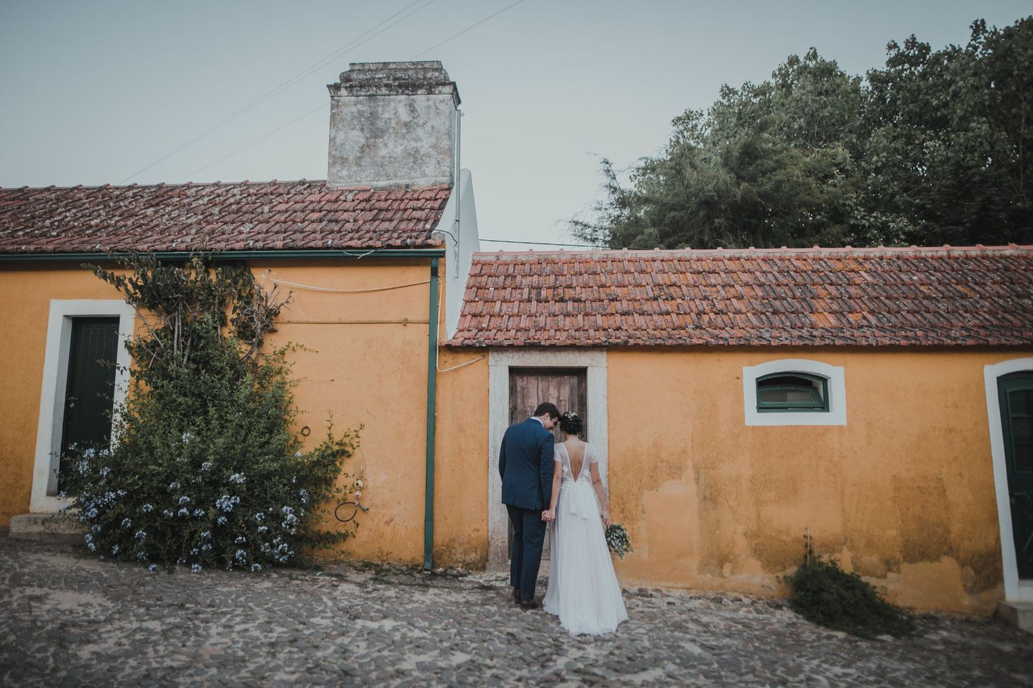 Wedding couple posing against yellow walls of rustic building with terracotta roof.