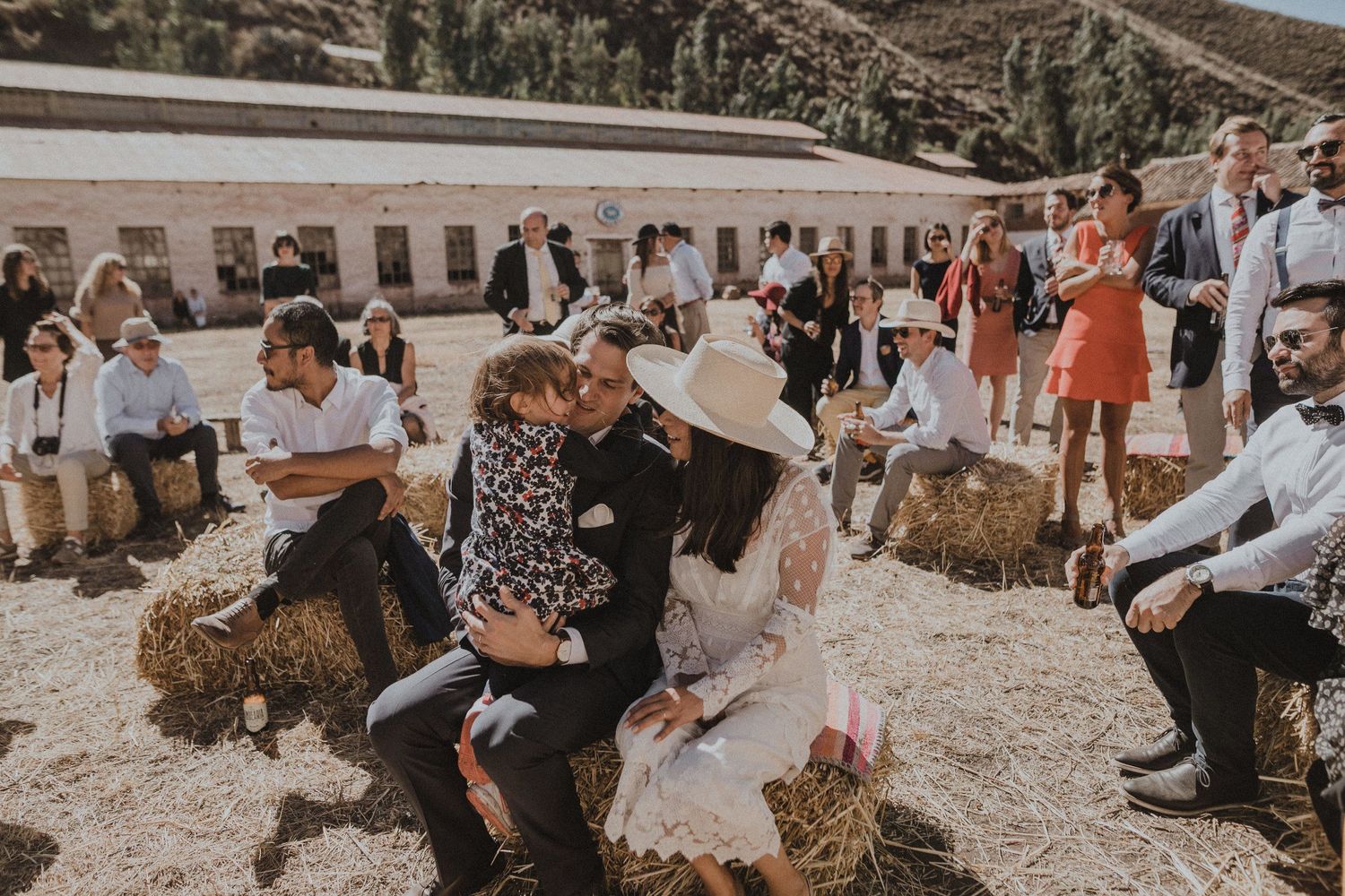 Wedding party relaxes on hay bales during outdoor rustic celebration.
