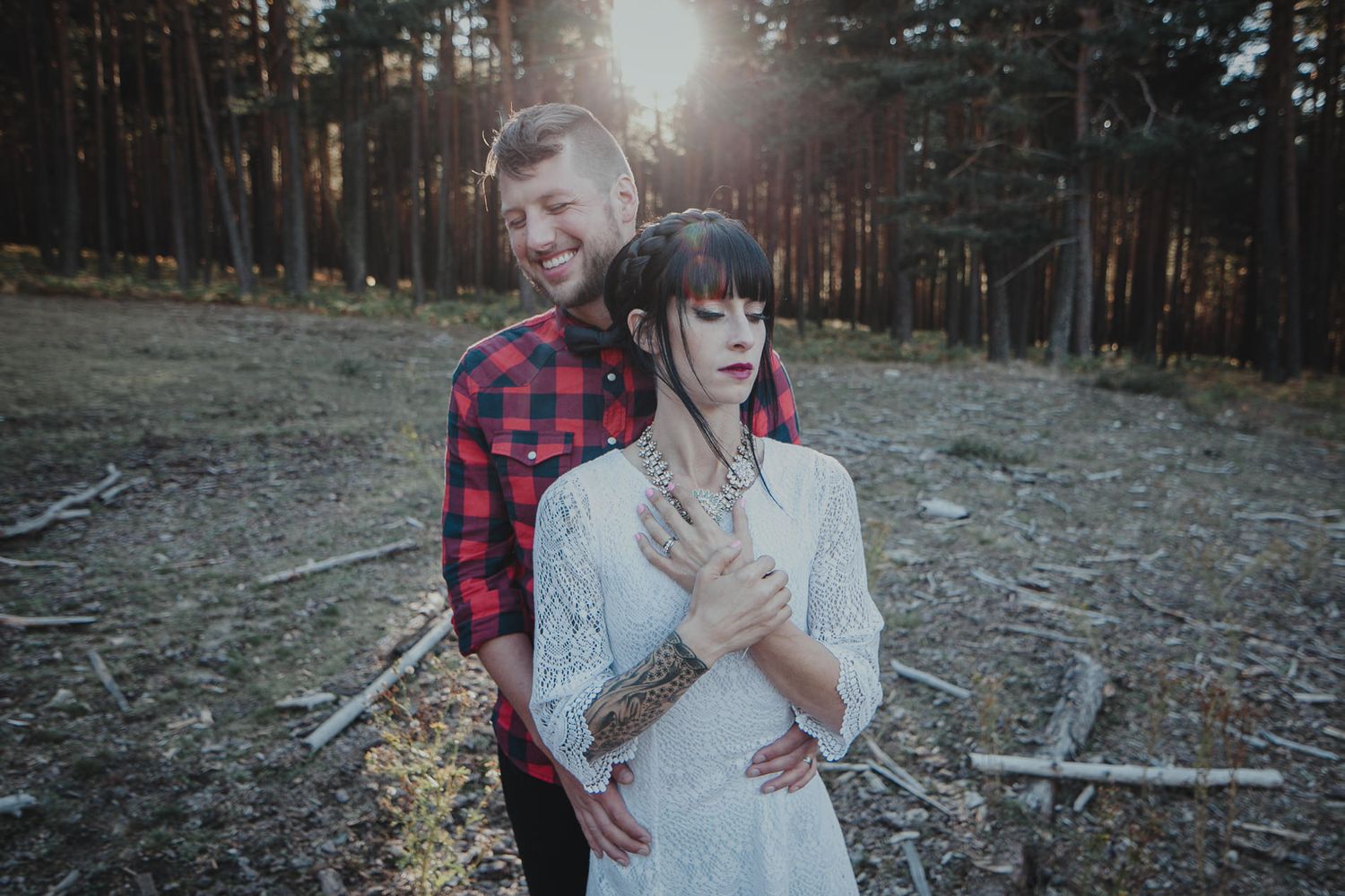 Couple poses in sunlit forest clearing wearing red plaid shirt and white lace dress.
