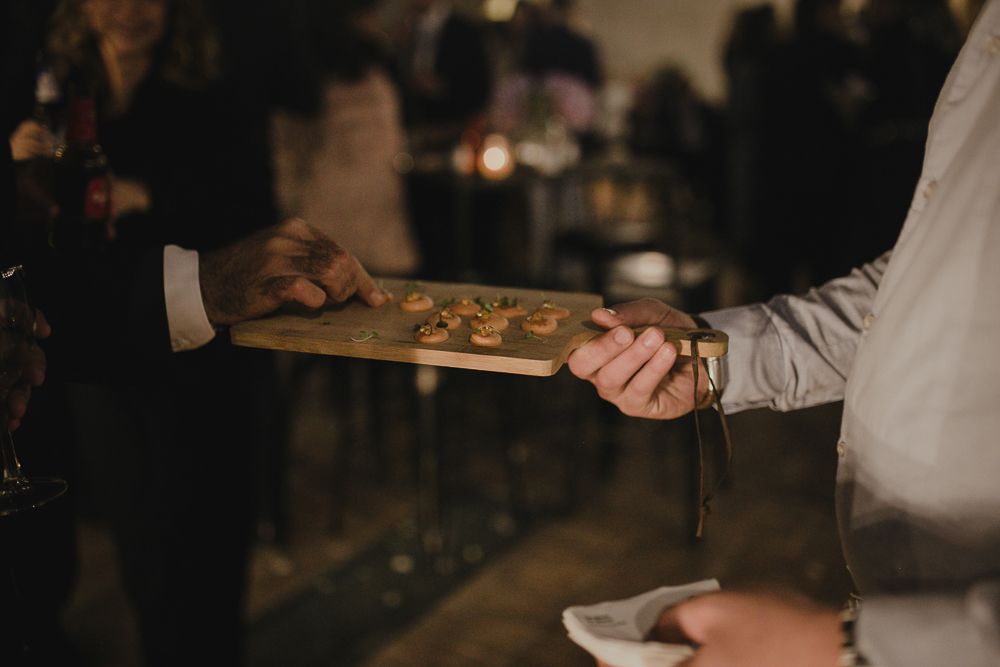 Server presenting appetizers on a wooden board at an elegant event in dim lighting.