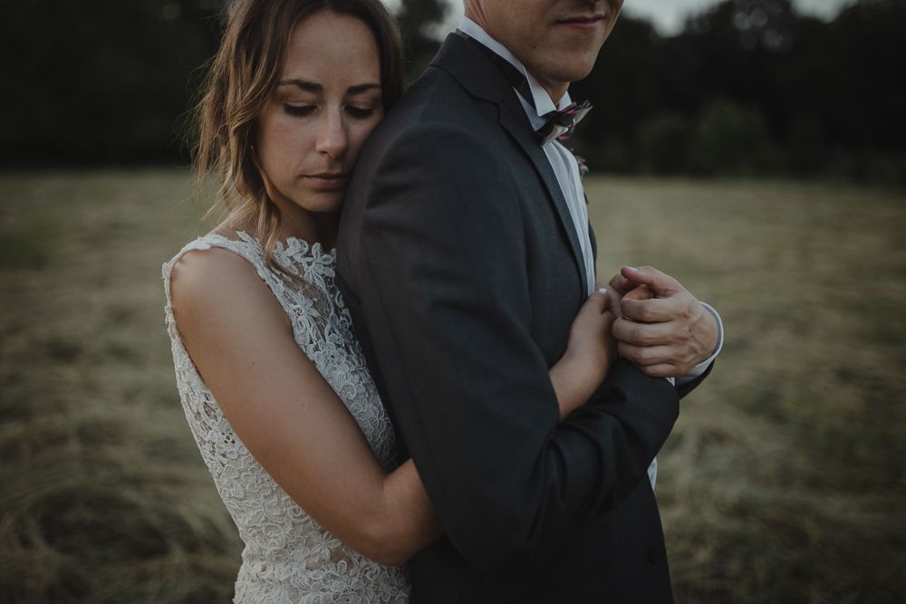 A romantic wedding portrait in a moody field captures the couple embracing in formal attire at dusk.