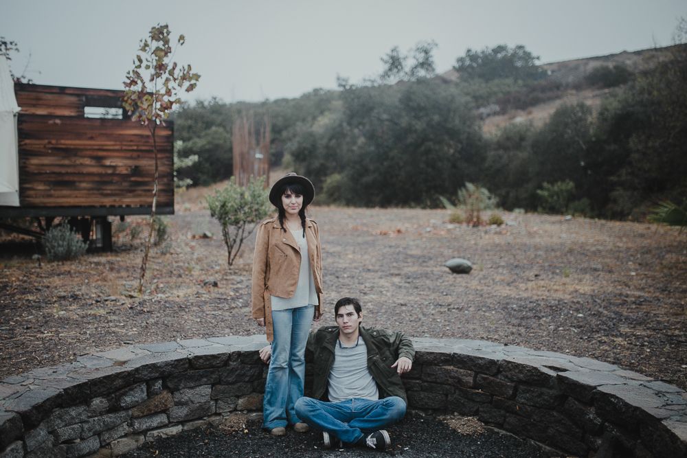 A casual couple poses in a rustic outdoor setting with mountains in the background.