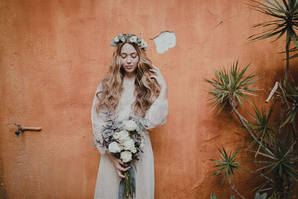 A bohemian bride in a lace dress with floral crown poses against a terracotta wall with desert plants.