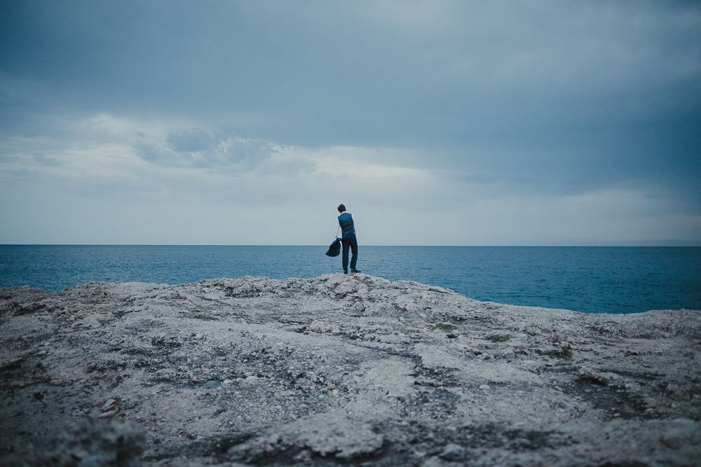 A silhouetted figure stands alone on a rocky ocean cliff against a blue sky.