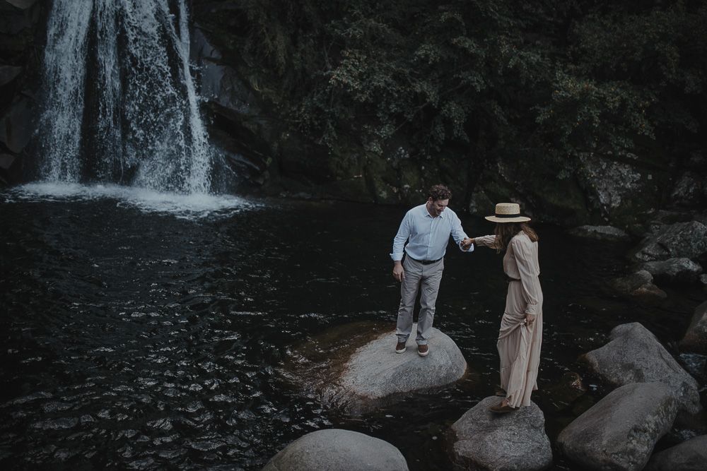 A couple stands on rocks near a cascading waterfall in a moody nature setting.