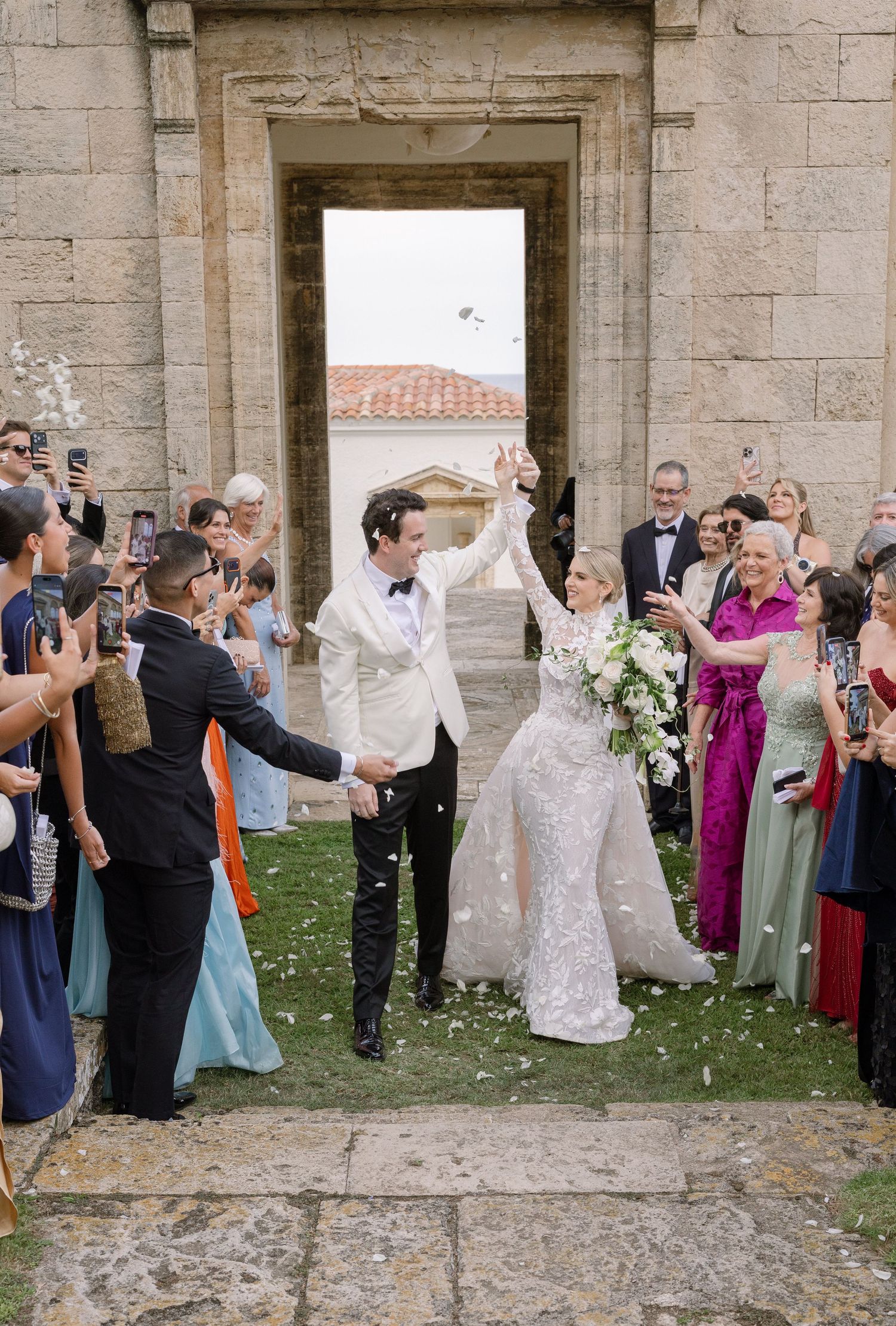 Bride and groom celebrate wedding exit through stone church doorway as guests toss white flower petals.