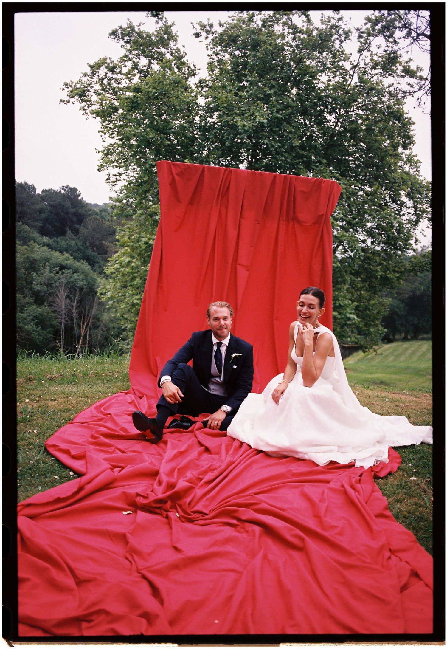 A couple sits on a large red blanket spread out on grass during an outdoor photo session.