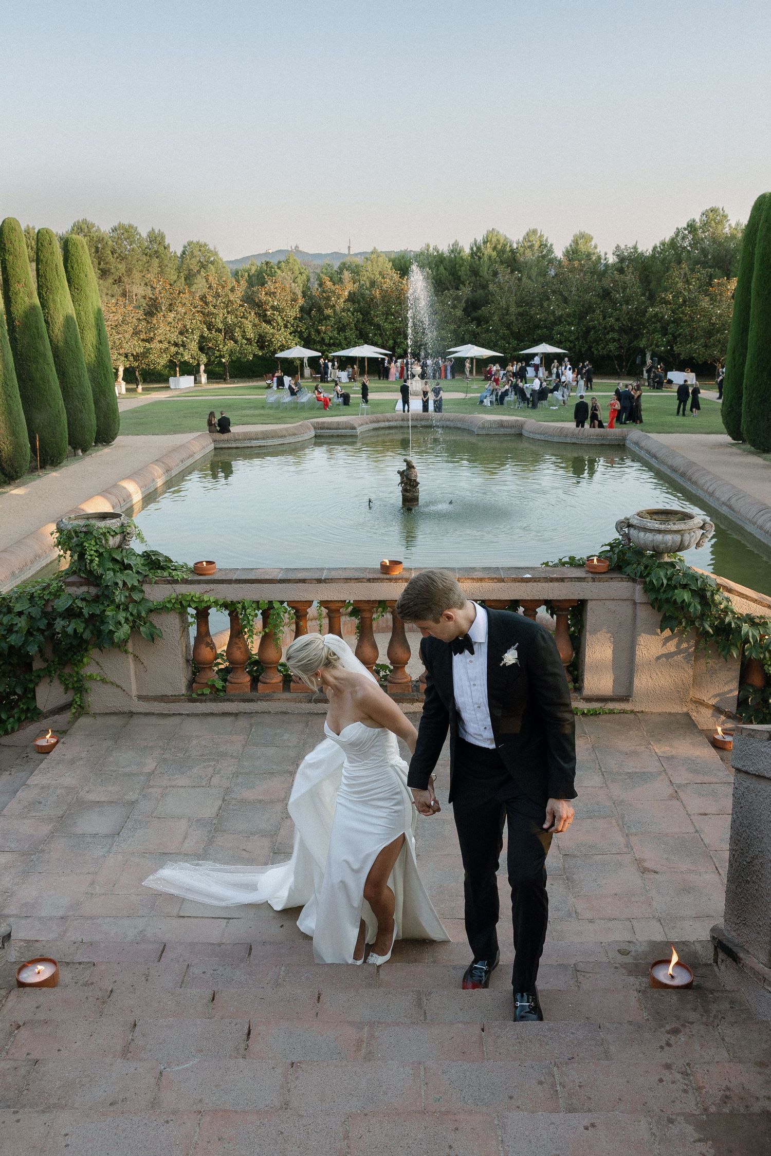 Couple walks along stone terrace with fountain and garden views at sunset.