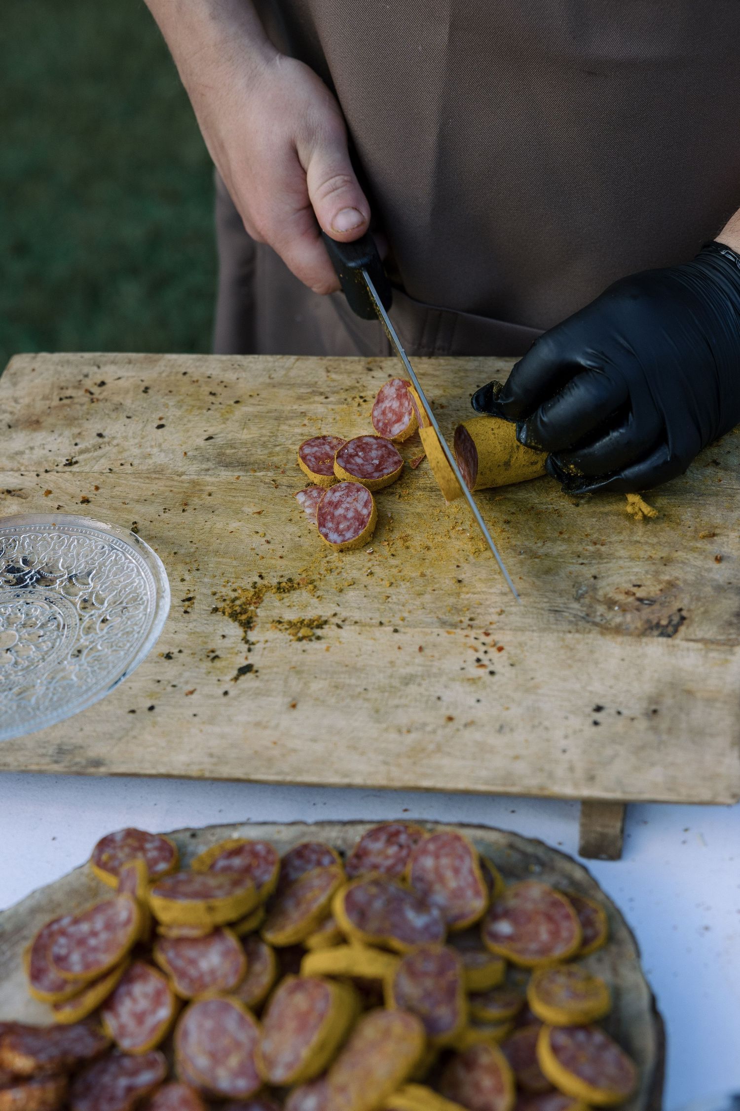 Hands in black gloves slicing cured meats on a wooden cutting board at a catering station.