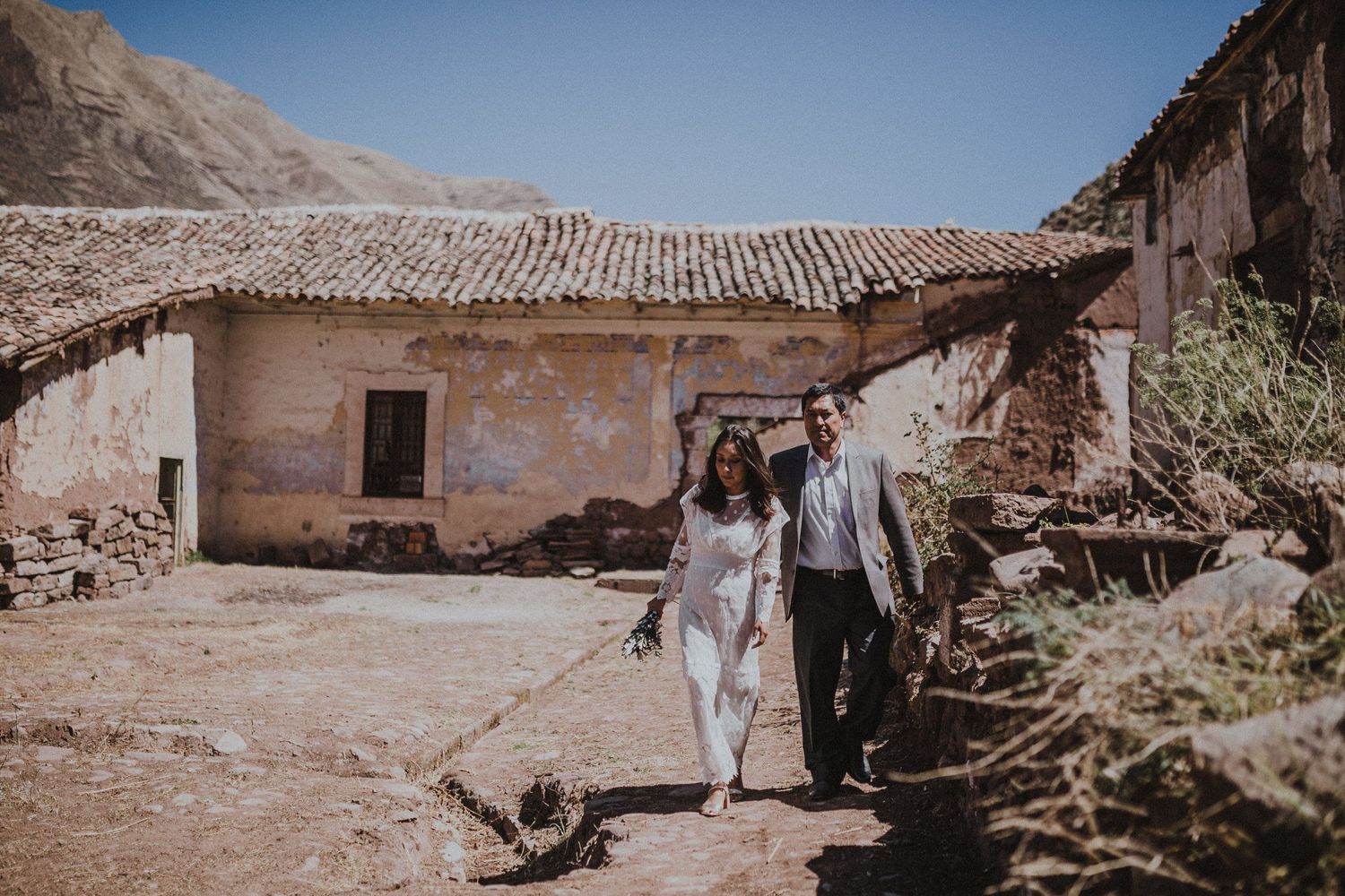 A wedding couple walking along a rustic path near an old adobe building in a desert setting.