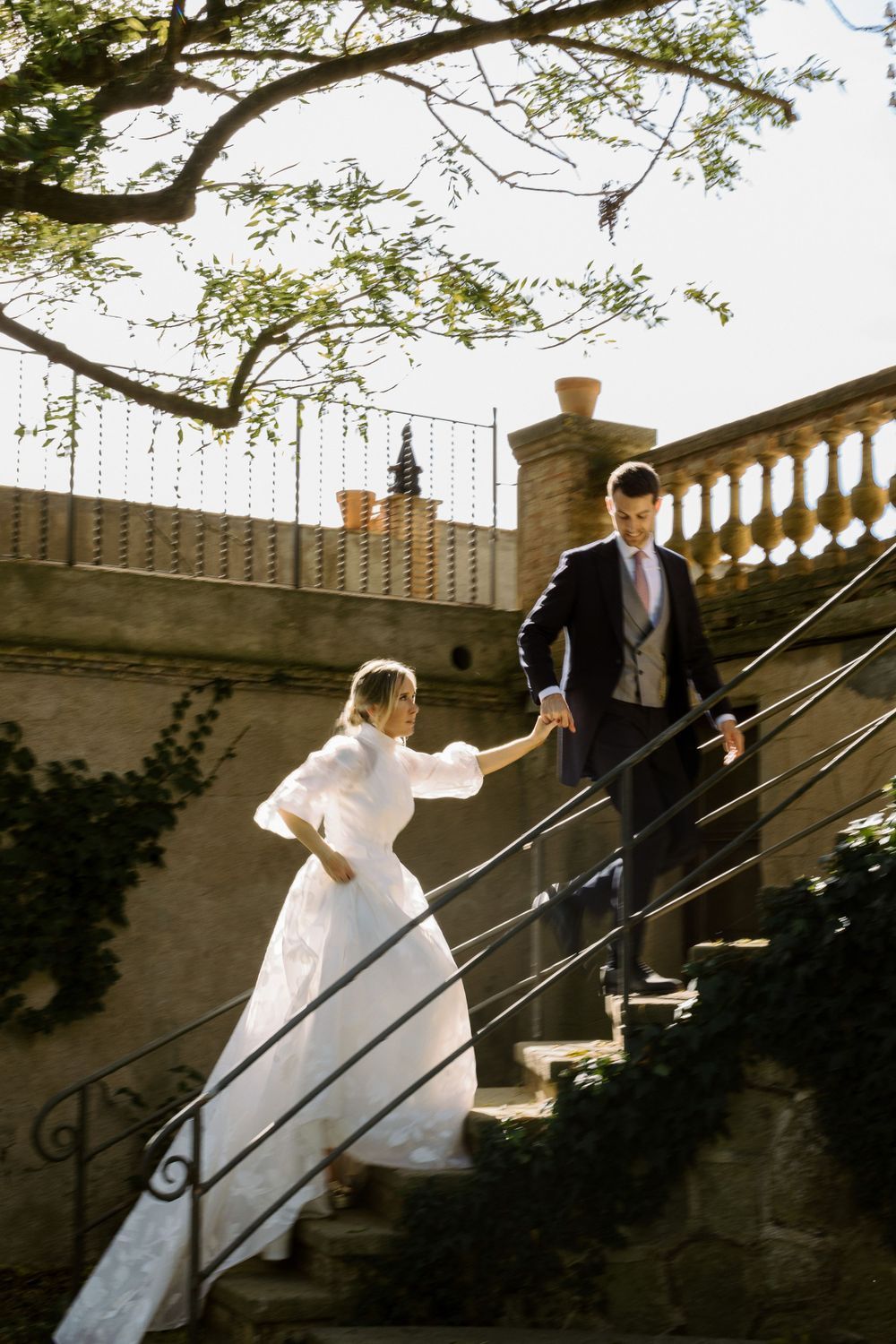 A couple in wedding attire hold hands while descending ornate stone stairs.