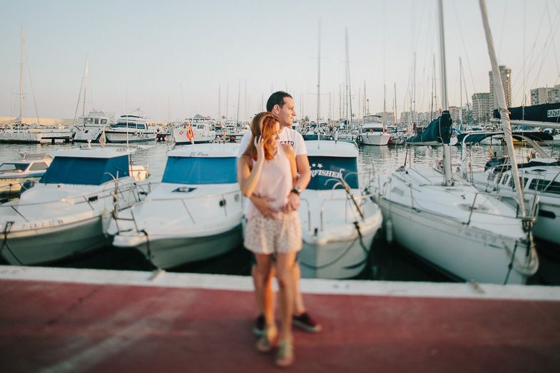 A couple shares a romantic moment at a marina with boats docked in the background at sunset.