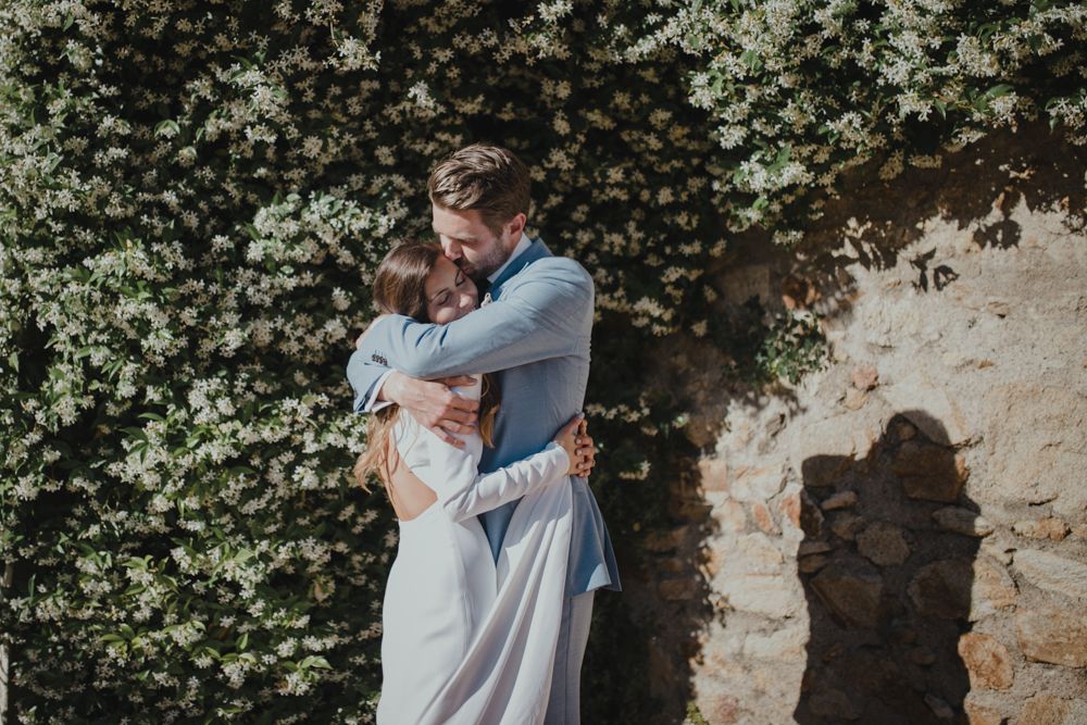 Romantic outdoor portrait of newlyweds embracing against stone wall with flowering vines.