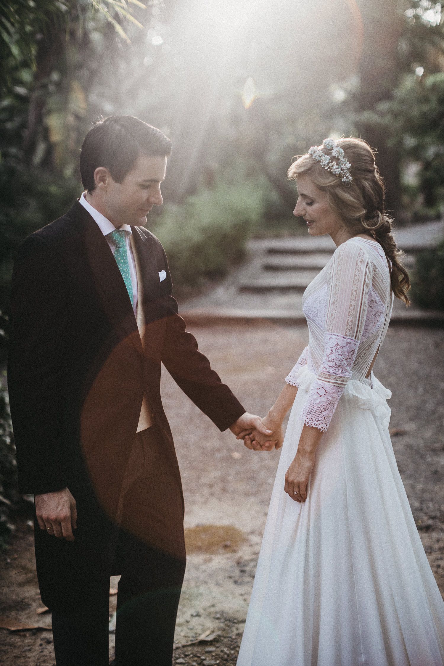 Sunlit wedding portrait of couple in formal attire holding hands on garden steps.