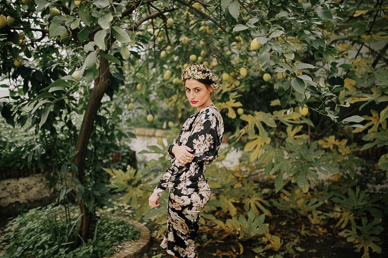 Woman in floral dress poses elegantly among green foliage and trees.