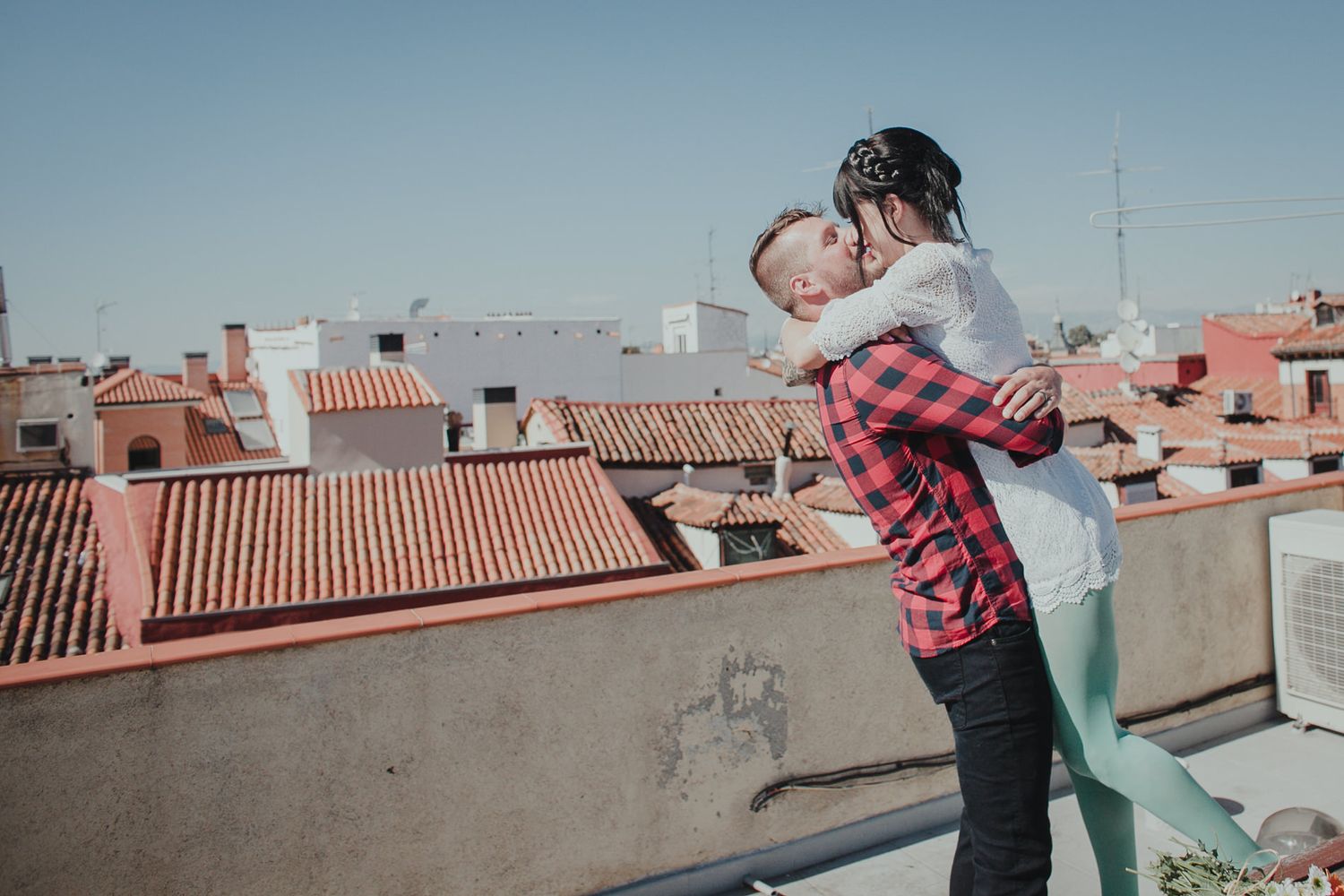 Romantic couple embraces on a rooftop terrace overlooking terracotta tiles and urban buildings against blue sky.