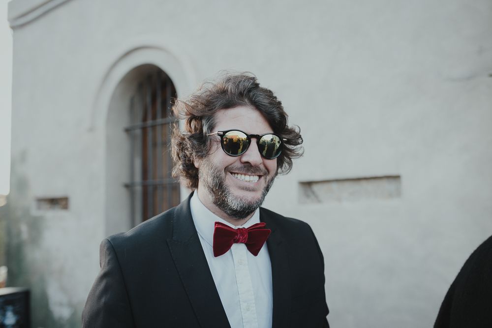 Wedding guest in black suit and bowtie smiling outdoors.