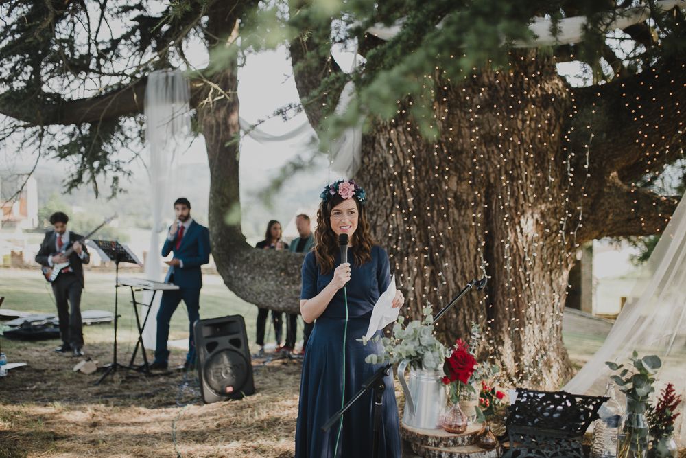 Musicians perform at an outdoor ceremony under tall evergreen trees with fairy lights.