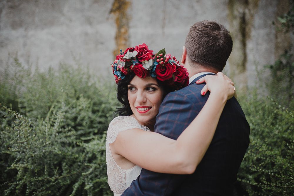 A beautiful bride wearing a red flower crown embraces her partner in a garden setting.