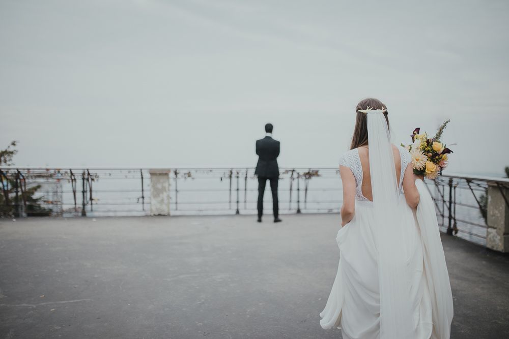Couple in wedding attire standing on an overcast deck overlooking water with railings.
