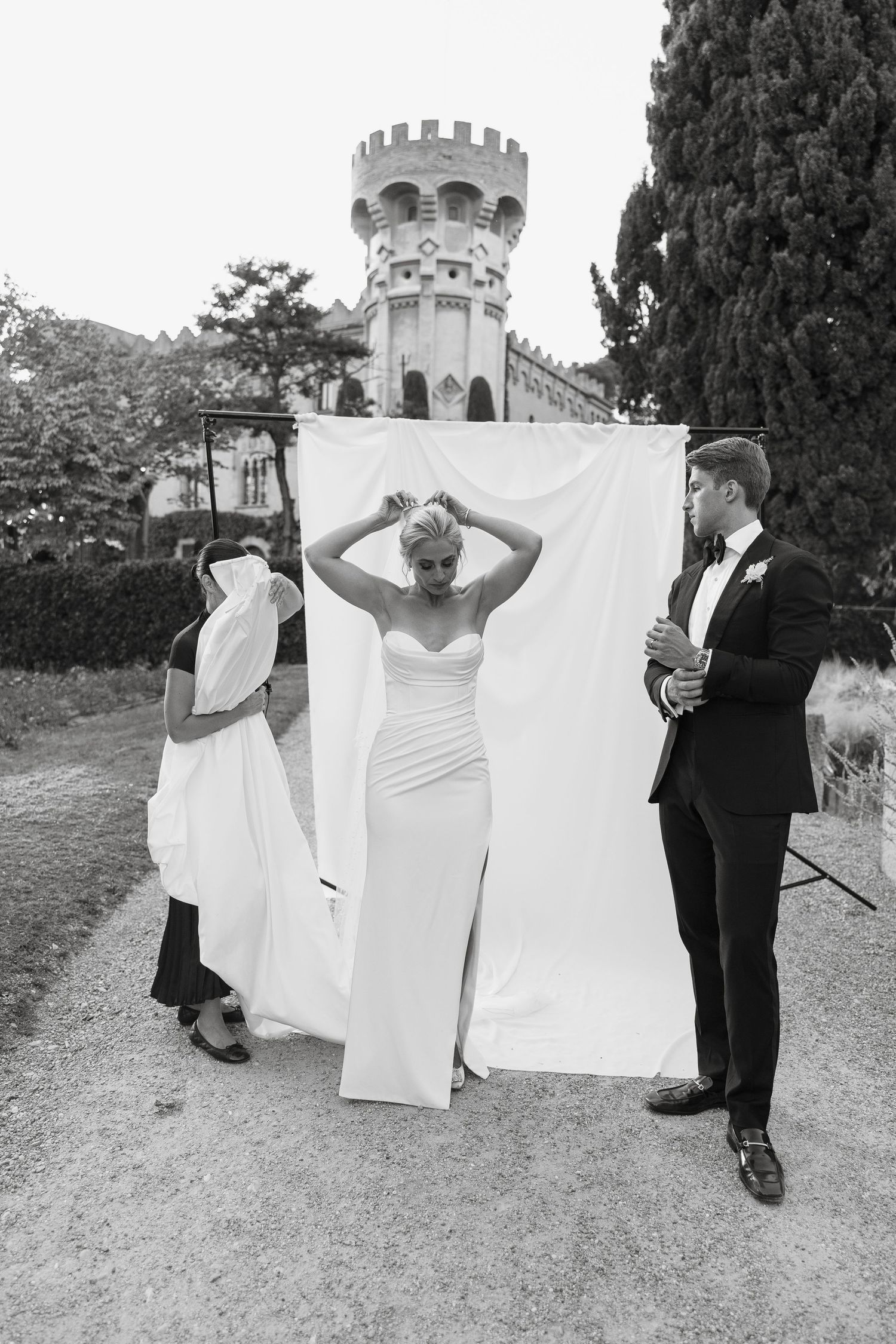 A wedding party poses playfully behind white sheets hanging outdoors near a castle tower.