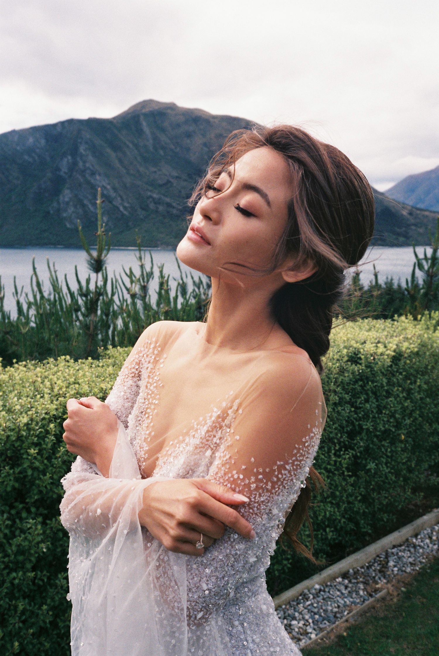 A person in a delicate white lace dress poses against a scenic mountain lake backdrop with wildflowers in the foreground.