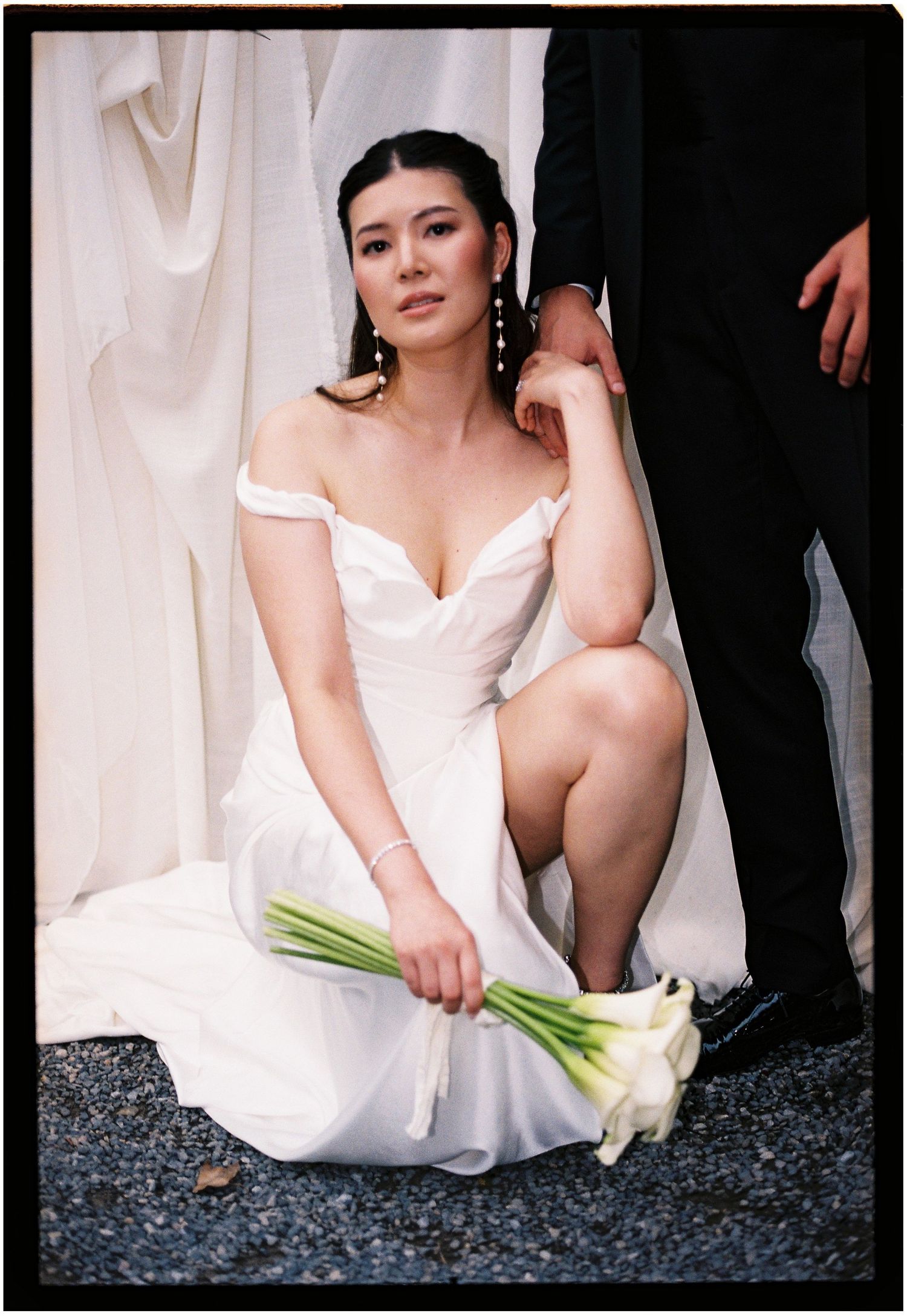 A figure in a white dress poses elegantly on dark stones while holding calla lily flowers.