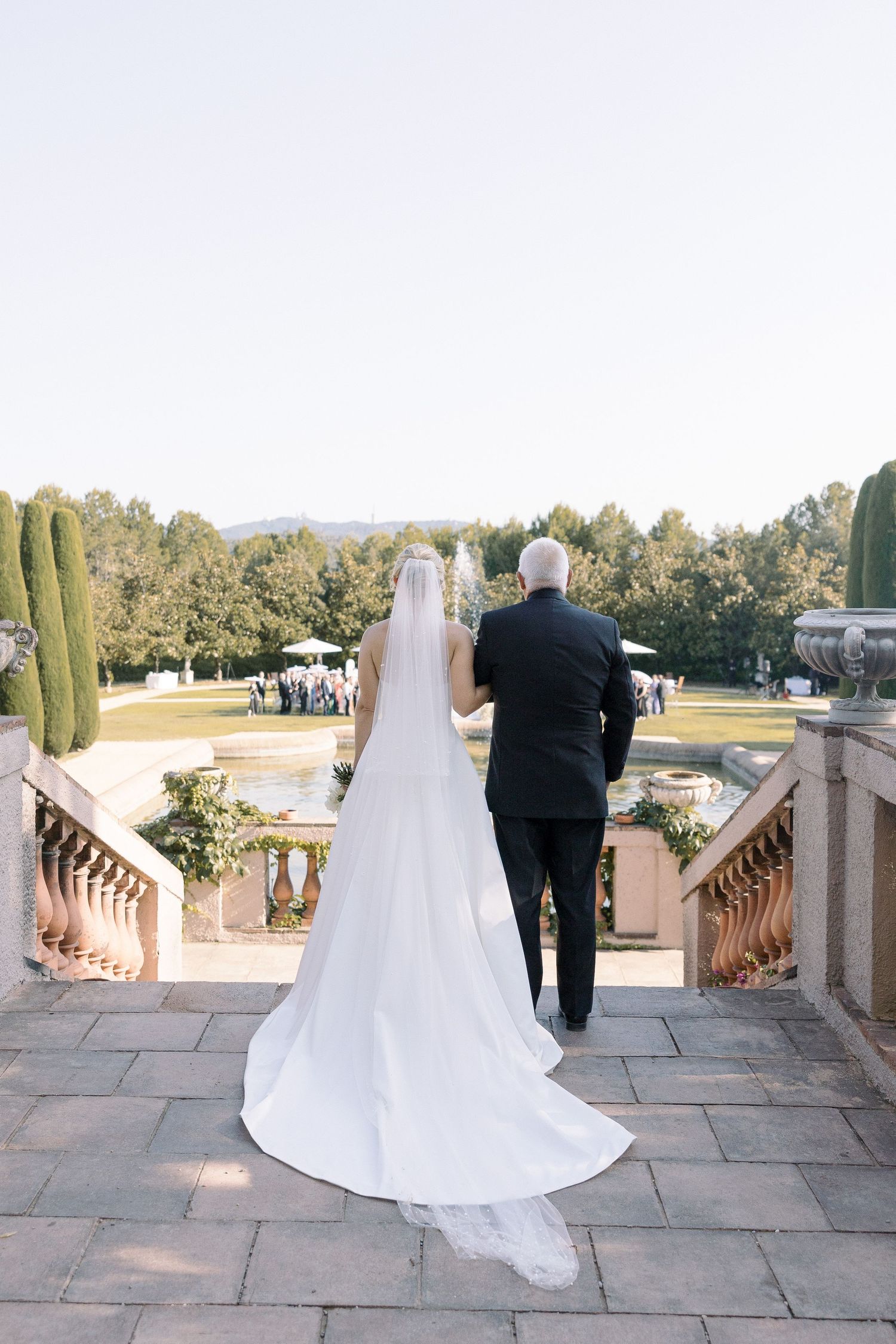 A long white wedding dress train trails down stone steps overlooking a formal garden.