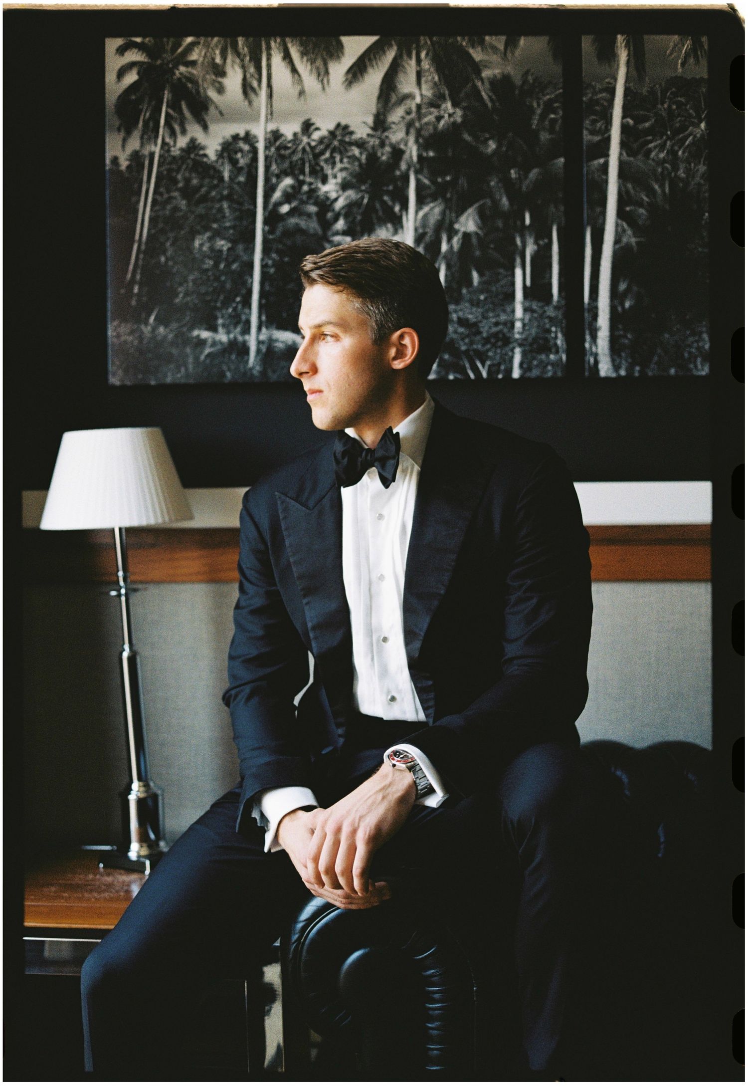 Groom in classic tuxedo sitting in room with tropical palm tree artwork.