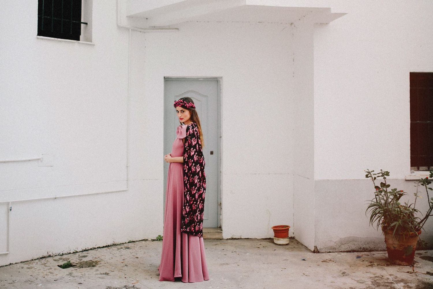 Someone wearing a long pink dress with floral print kimono stands in doorway of white Mediterranean building exterior.
