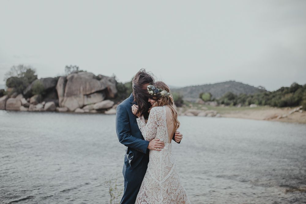 Elegant wedding couple embraces on shoreline with dramatic rock formations in background.