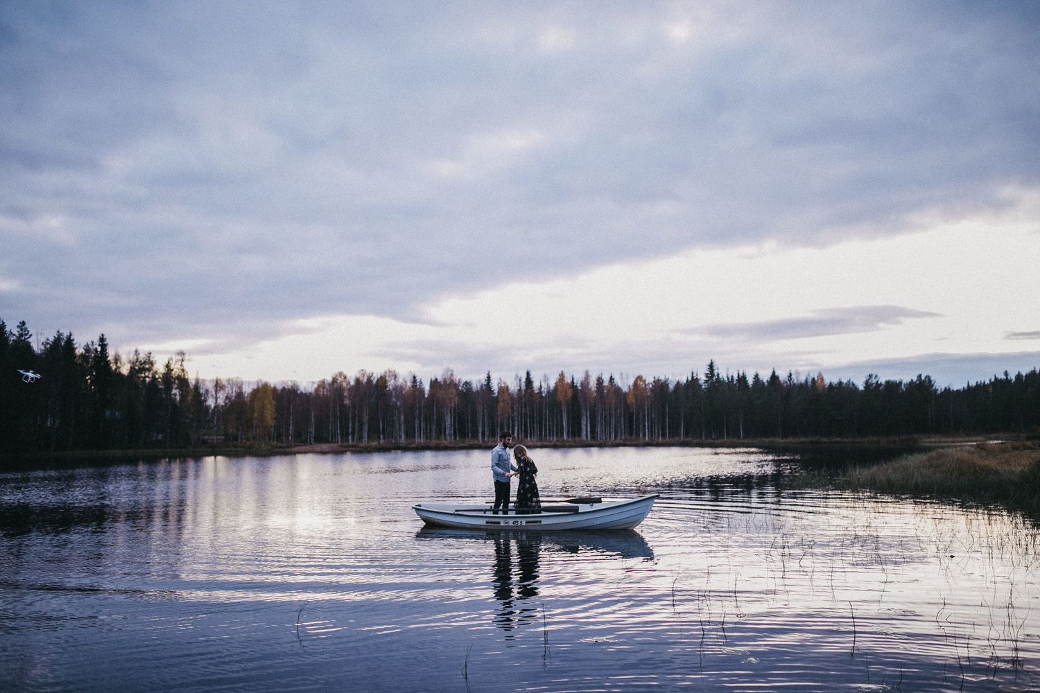 Silhouettes of a couple standing in a boat on a tranquil lake surrounded by pine trees at dusk.