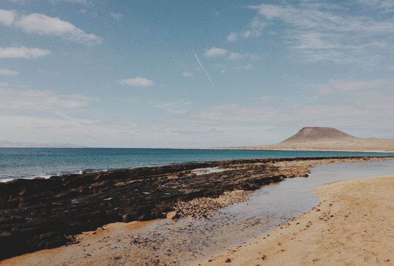 Rocky coastline with volcanic black rocks and sandy beach leading to a distant mountain under blue sky.