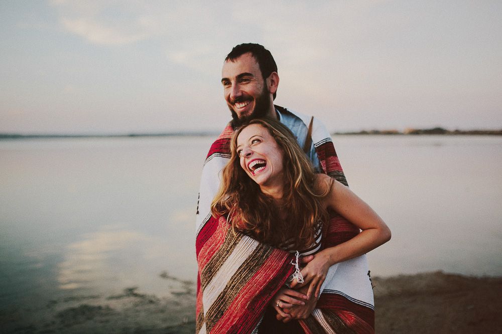 Couple sharing laughter while wrapped in striped blanket by the water.
