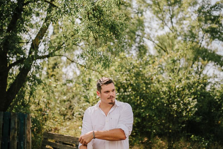 Person in white shirt stands thoughtfully in outdoor garden setting with sunlit trees.