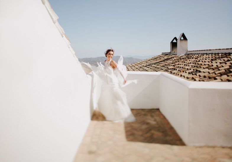 A white wedding dress billows in the wind against white Mediterranean architecture and terracotta roof tiles.