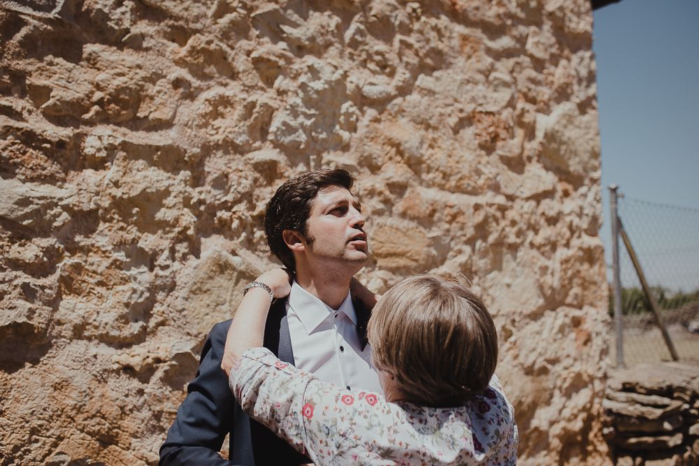 Two figures embrace against a rustic stone wall backdrop in warm sunlight.