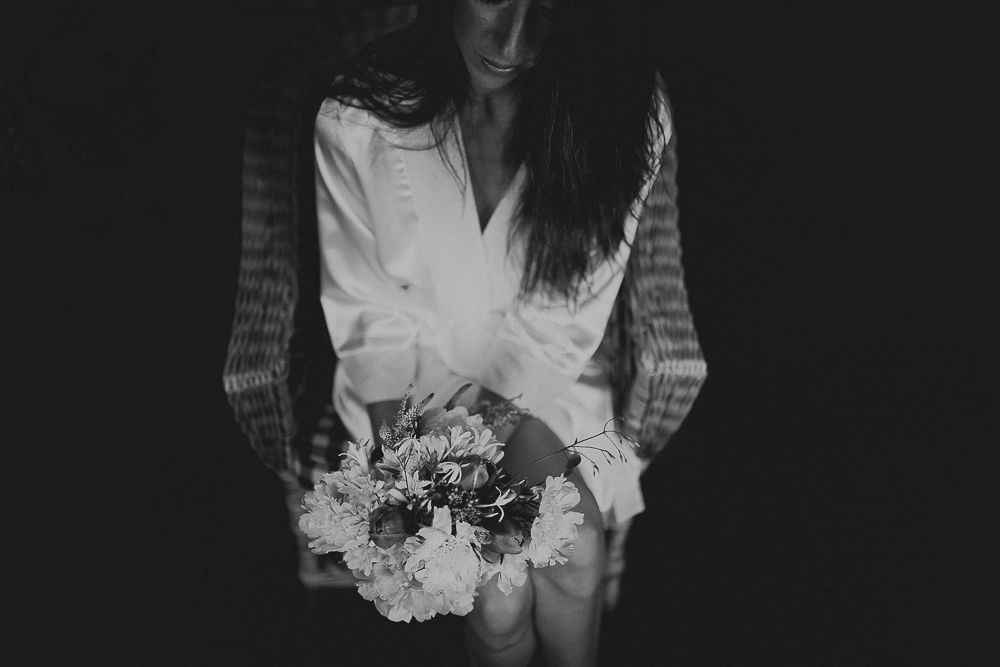 Person in white robe holding a bouquet of flowers in dramatic black and white lighting.