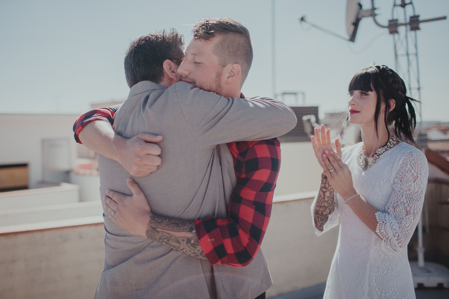 Two people share an emotional embrace on a rooftop while another looks on in a joyful moment.
