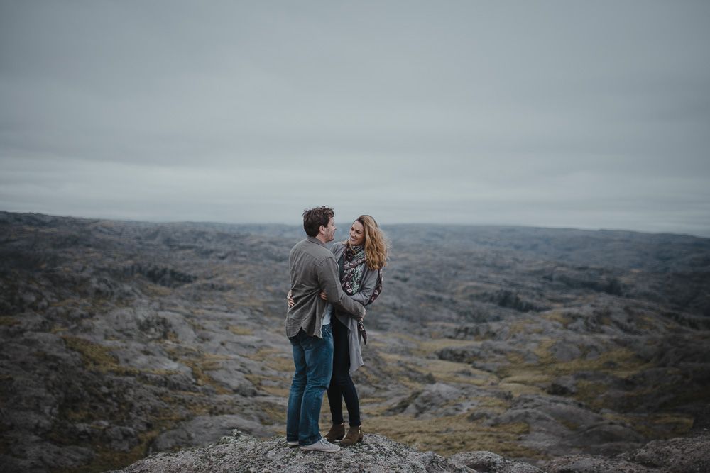 Two people share an intimate moment while standing on rocky terrain under cloudy skies.