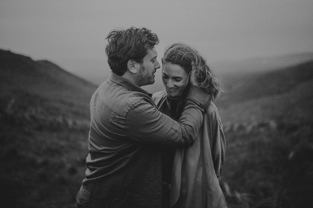 Couple shares tender moment against dramatic mountainous backdrop in black and white.