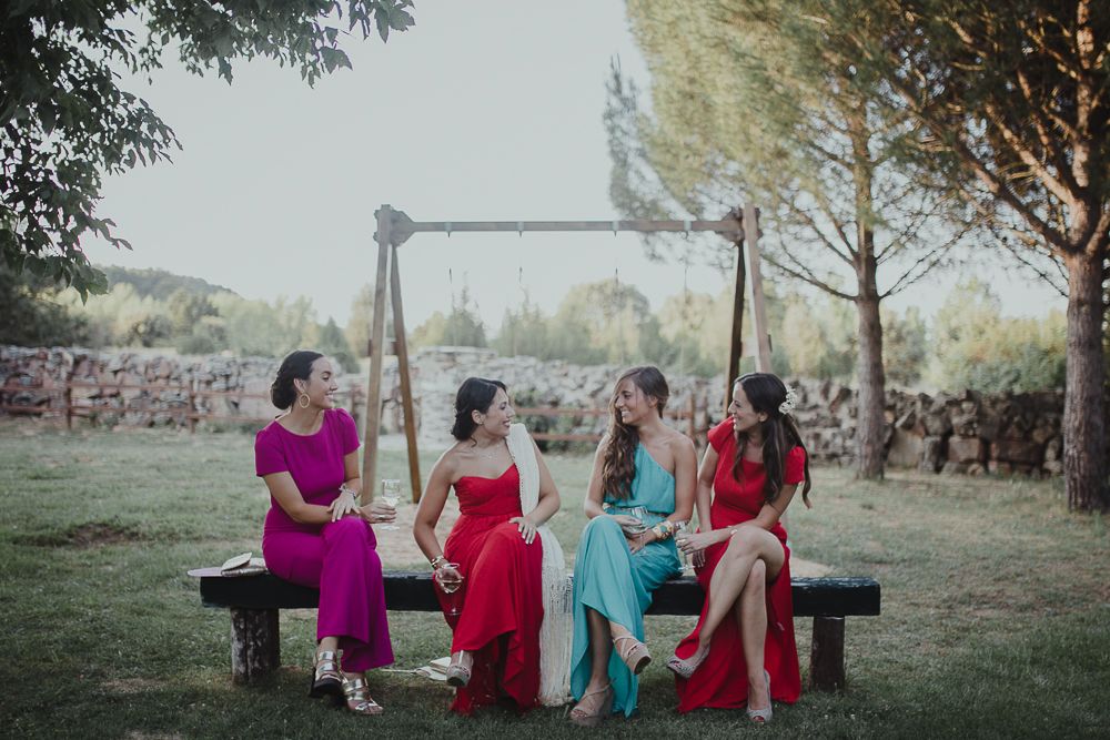 Friends in vibrant formal dresses sitting together on a rustic wooden bench outdoors.