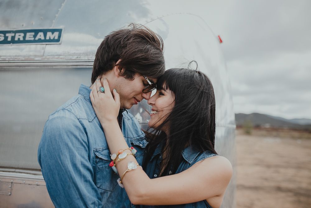 Two people share an intimate moment next to a silver Airstream trailer in the desert.